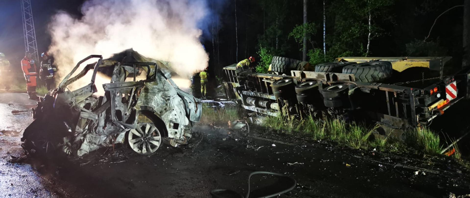 Zdjęcie przedstawia działania służb ratowniczych po wypadku i pożarze na drodze wojewódzkiej koło Wierchlesia. Zdjęcie wykonane w porze nocnej.