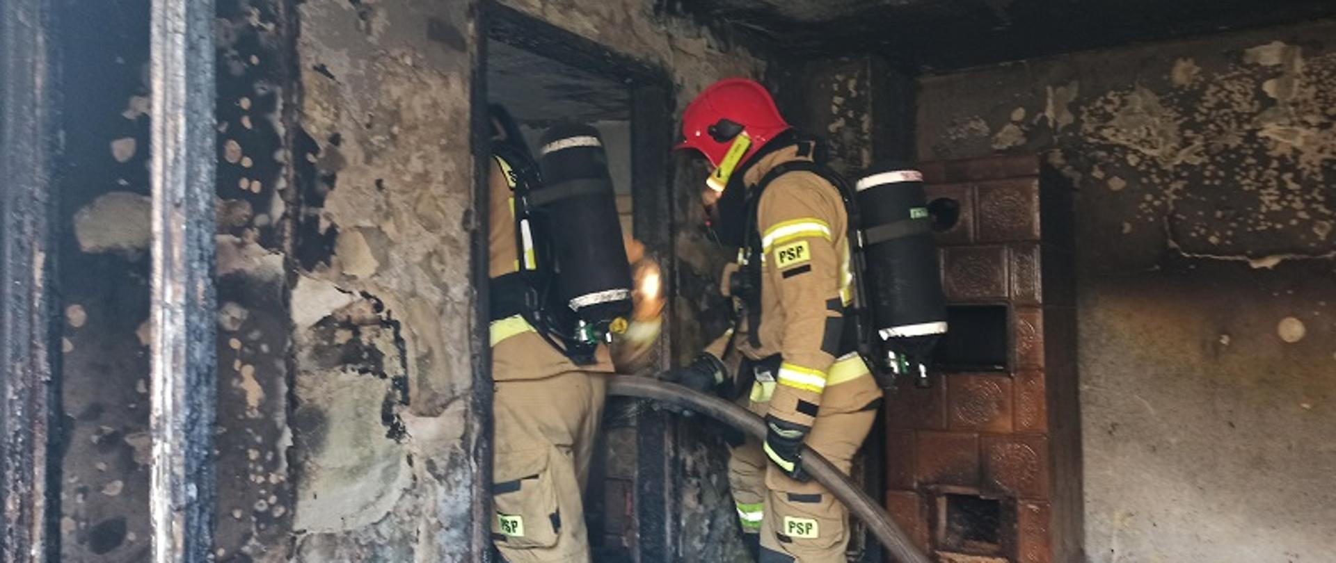 Zdjęcie przedstawia spalone wnętrze pomieszczenia i rotę strażaków z wężem gaśniczym w ubraniach specjalnych stojących we wnęce drzwiowej. Ściany z ubytkami tynku, czarne od ognia