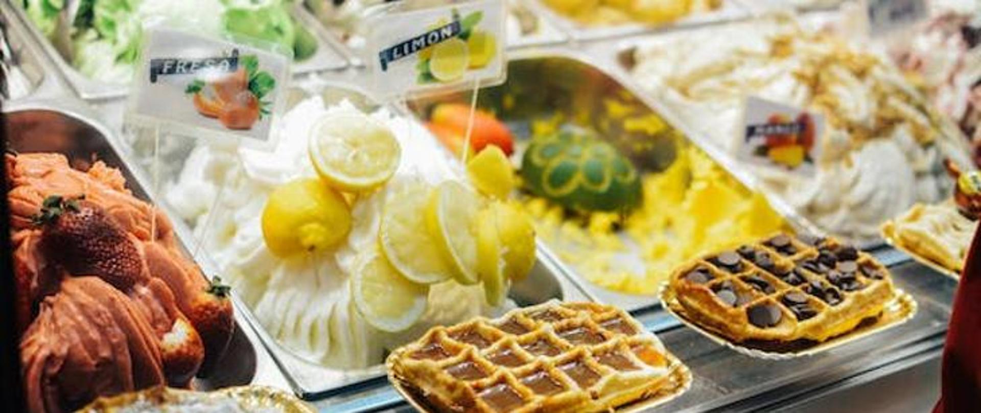 lody w pojemnikach w lodziarni i dwa gofry z czekolada na tackach jednorazowych na ladzie z lodami