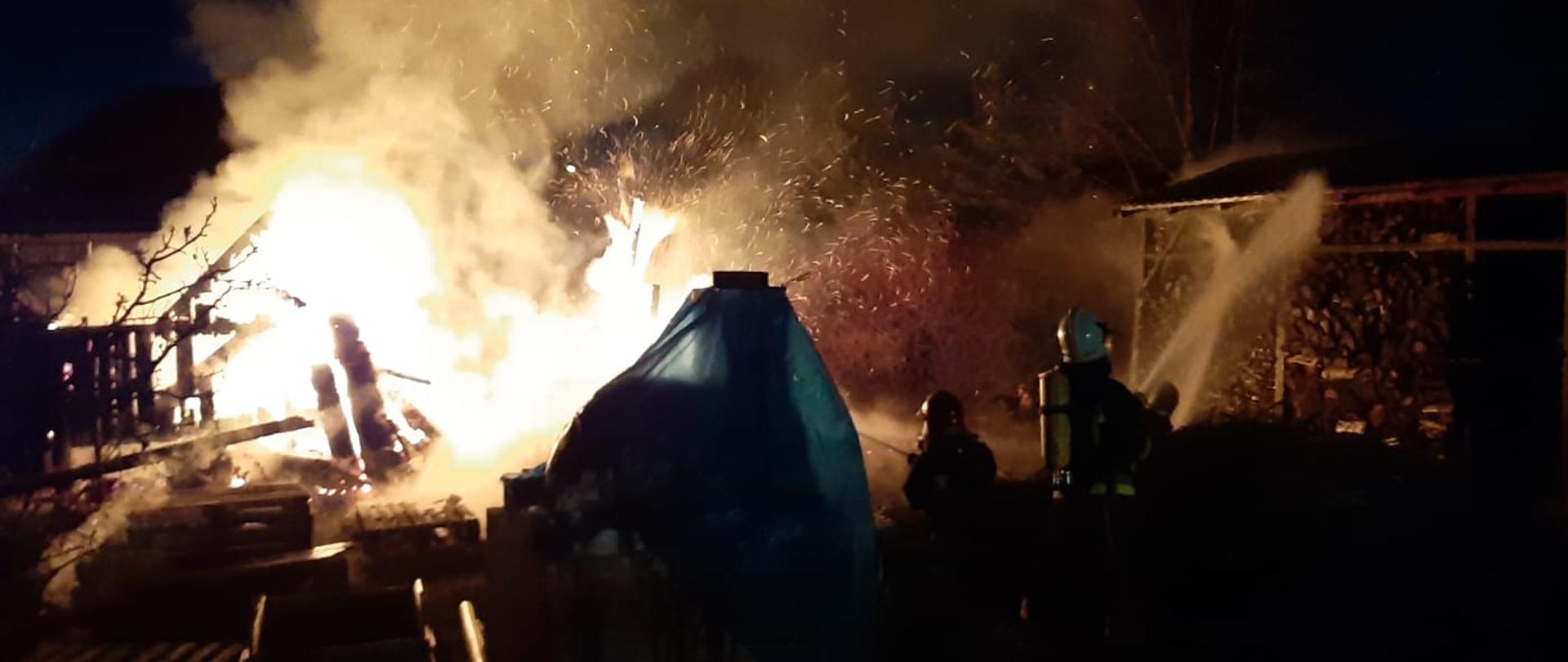 na zdjęciu strażacy podczas działań gaśniczych podający prądy wody na palący się drewniany domek i wiatę z drewnem