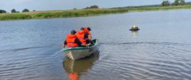 Ratownicy ubrani w kamizelki podpływają łodzią do osoby poszkodowanej. Osoba która pozoruję osobę poszkodowaną ubrana jest w kombinezon wypornościowy i kamizelkę unosząc się na rzece