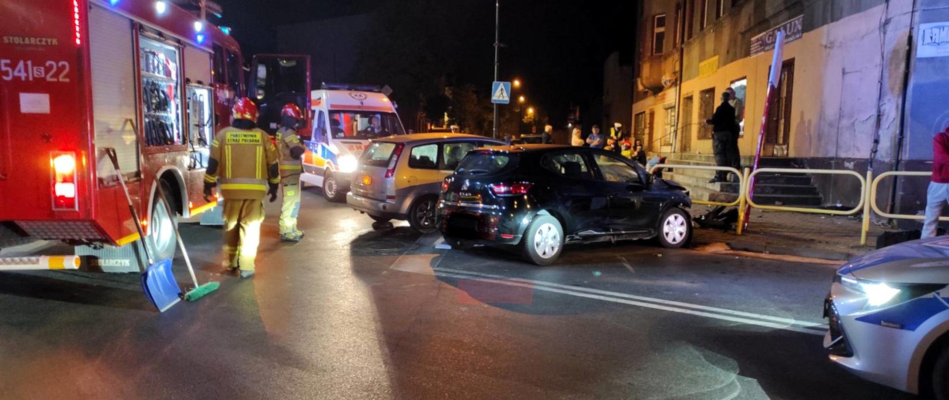 Zdjęcie przedstawia dwa rozbite samochody. Obok stoi średni samochód ratowniczo - gaśniczy, karetka Państwowego Ratownictwa Medycznego oraz radiowóz policji.
