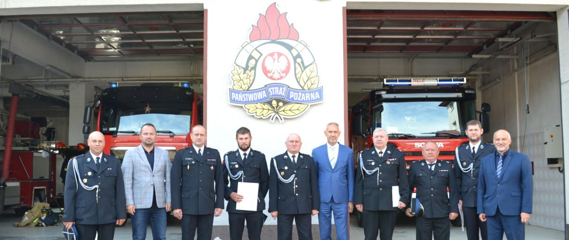 Na zdjęciu widać druhów OSP oraz komendanta powiatowego PSP z Ustrzyk Dolnych w mundurach galowych, prezesa WFOŚiGW oraz starostę powiatu bieszczadzkiego wraz z zastępcą. W tle garaże jednostki ratowniczo gaśniczej w których stoją wozy bojowe, oraz widniejące na ścianie logo PSP.