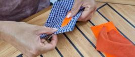 A woman’s hands sewing at a table, with scissors, a “Blue Door” sticker, and two fabric scraps — one orange and one green — placed nearby.