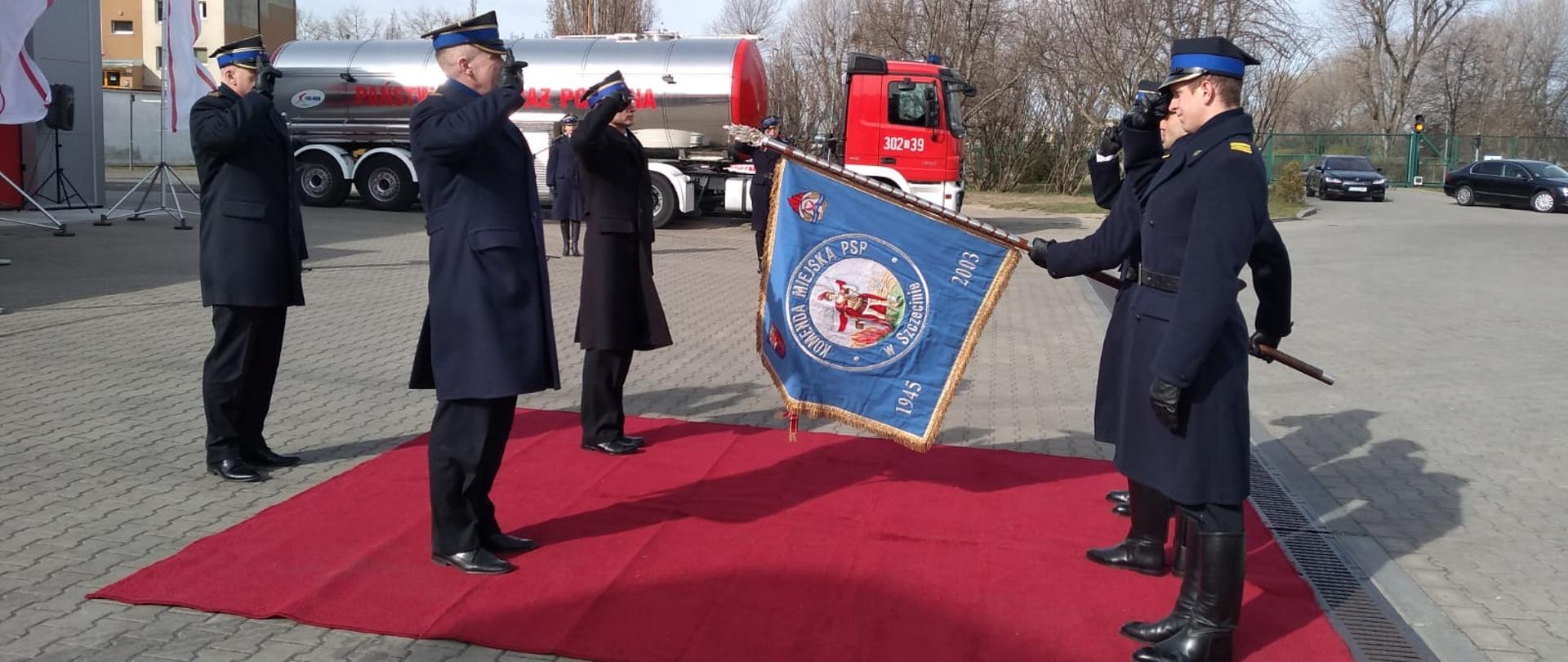 Na zdjęciu widoczna ceremonia przekazania obowiązków Komendanta Miejskiego PSP w Szczecinie. Na zdjęciu widoczni po lewej sronie komendanci, po prawej zaś poczet sztandarowy z opuszczonym sztandarem.
W tle widoczna cysterna. Strażacy salutują. Przekazanie obowiązków odbywa się na czerwonym dywanie