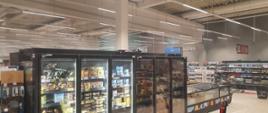 Zdjęcie wewnątrz sklepu. Widać półki sklepowe. Na pólkach produkty. Jedna z lodówek okopcona od dymu. Oświetlenie sztuczne.