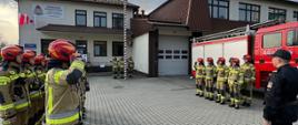 Na zdjęciu widoczny na placu wewnętrznym przed Komendą PSP w Gorlicach poczet flagowy przy maszcie w ubraniach specjalnych w kolorze piaskowym i czerwonych hełmach podczas podnoszenia flagi w zawiązku z uroczystością Dnia Flagi Rzeczypospolitej Polskiej. Przed pocztem ustawione zmiany służbowe w dwuszeregu w pozycji na baczność również w ubraniach specjalnych koloru pisakowego i czerwonych hełmach.