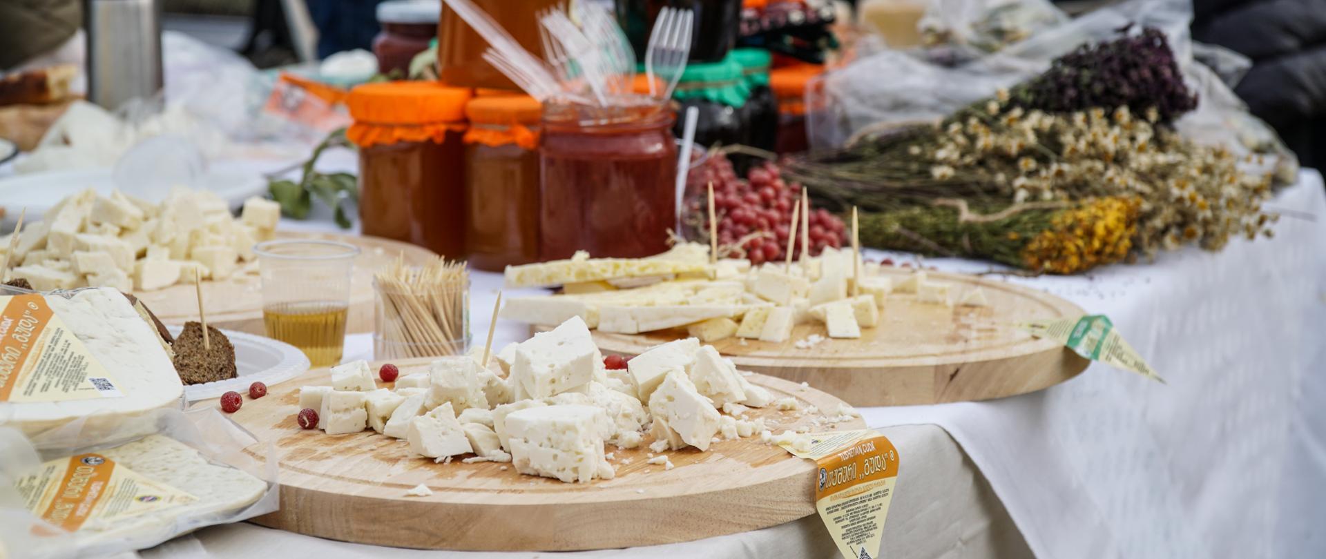 Kawałki sera leżą na tacach, w tle stoją słoiki i suszone kwiaty
