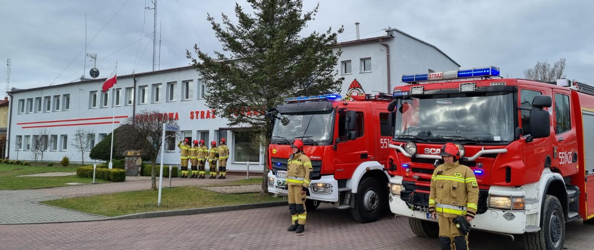 Zdjęcie przedstawia zmianę służby Jednostki Ratowniczo-Gaśnicze, którzy znajdują się przy maszcie flagowym, ubrani w odzież specjalną z hełmami. W tle widać napis "Państwowa Straż Pożarna" na budynku komendy, drzewa. Na pierwszym tle znajdują się dwa wozy bojowe z kierowcami przed autami.