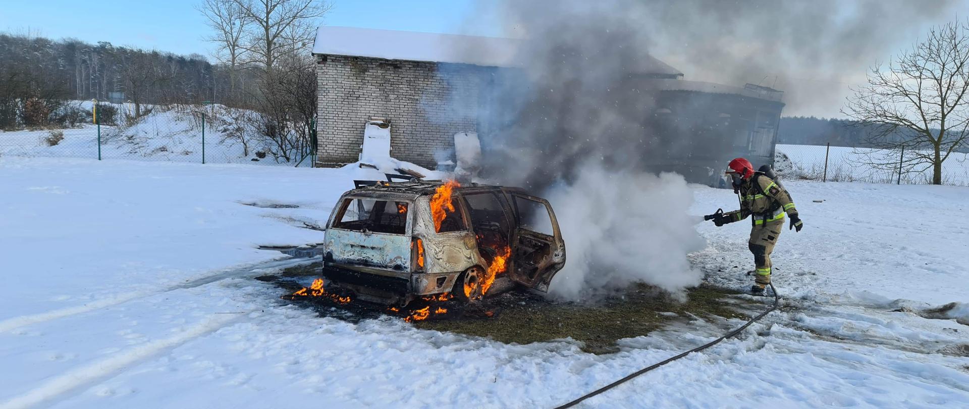 Na głównym planie widoczny palący się samochód osobowy, obok niego strażak ubrany w ubranie specjalne oraz aparat ochrony dróg oddechowych gaszący pożar, w tle widoczny budynek gospodarczy.