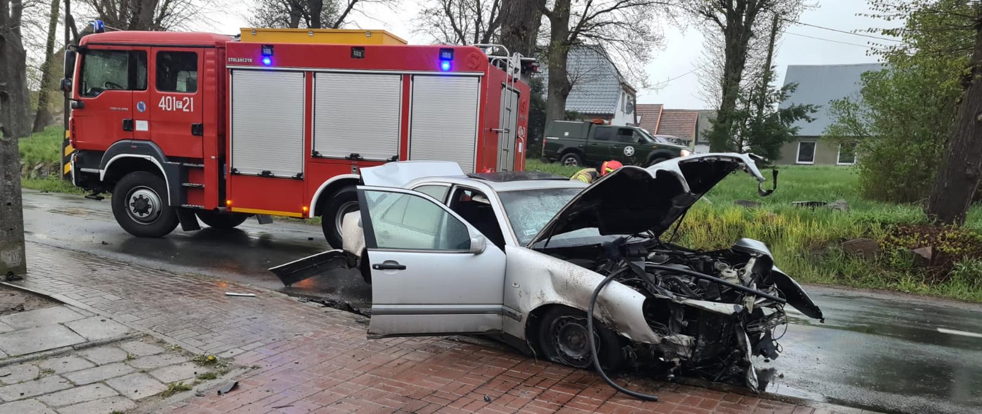 Fotografia przedstawia ujęcie prostopadłe do przebiegu drogi powiatowej. Na pierwszym planie w centralnej części kadru widoczny zniszczony samochód osobowy koloru srebrnego. Samochód stoi na pasie jezdni (widoczny przód i prawy bok auta) z otwartą klapą silnika, otwartą klapą tylną oraz otwartymi przednimi prawymi drzwiami. Widoczne są zniszczenia przedniego zderzaka, przedniego prawego koła i nadkola. Za samochodem, po lewej stronie fotografii na drugim pasie jezdni stoi lewym bokiem do fotografującego samochód gaśniczy, ma włączone światła ostrzegawcze. Dalej w tle widoczne zabudowania wiejskie oraz drzewa. Jest dzień, niebo jest zachmurzone, jezdnia jest mokra.