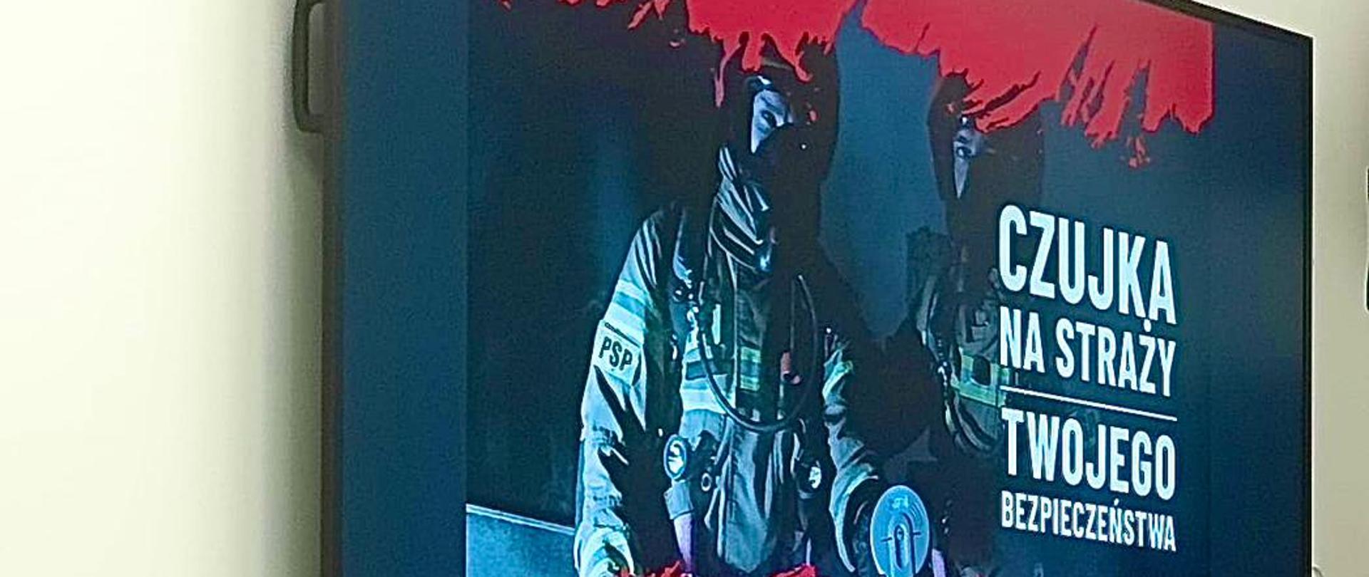 Na zdjęciu widać strażaka Państwowej Straży Pożarnej siedzącego przy stole podczas spotkania lub prezentacji. Ma na sobie mundur służbowy, a przed nim znajdują się dokumenty, mikrofon, filiżanki oraz laptop. Na ekranie za nim wyświetlona jest plansza z hasłem: „Czujka na straży Twojego bezpieczeństwa”. Grafika przedstawia strażaków w aparatach ochrony dróg oddechowych oraz element czujnika (najprawdopodobniej czujnika dymu lub tlenku węgla).
