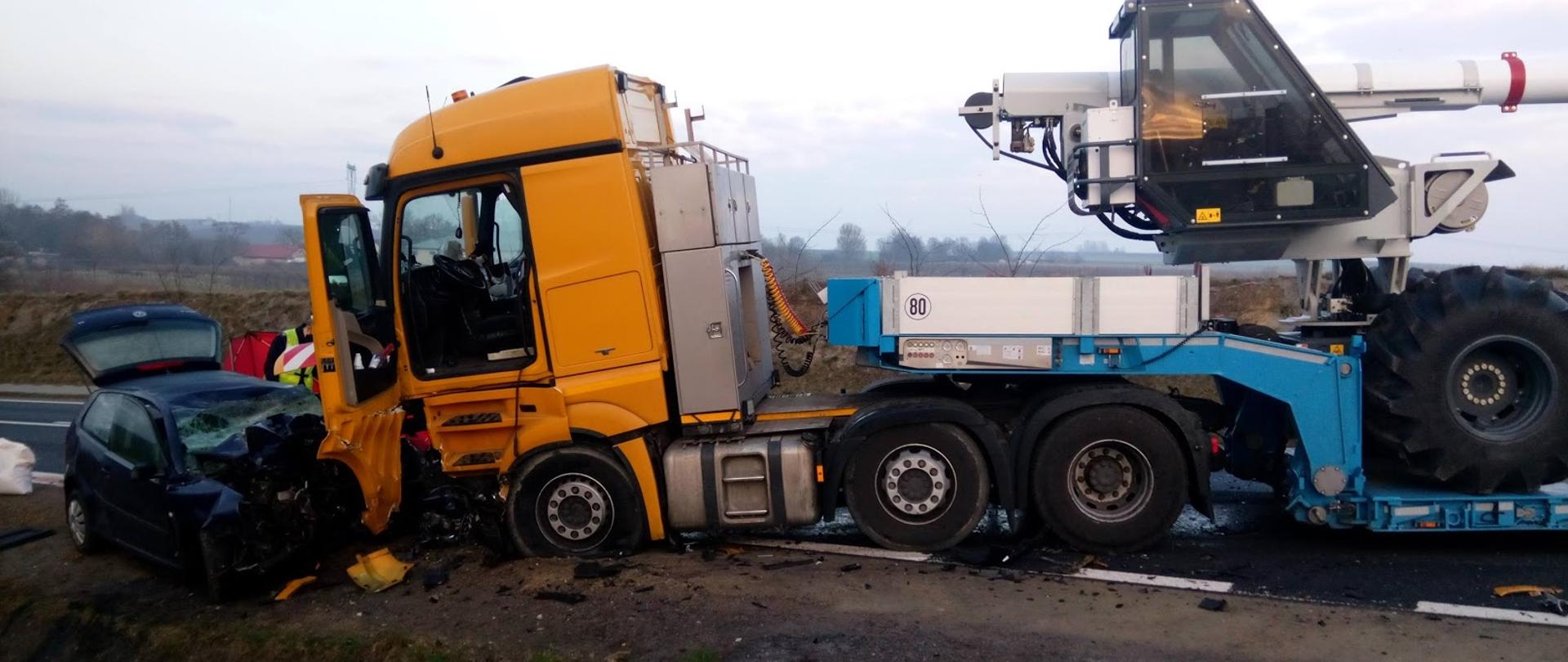 Samochód ciężarowy z otwartymi drzwiami kierowcy z przewożonym na naczepie sprzętem stojący na jednym pasie drogi poniżej zderzaka kabiny, samochód osobowy z widocznym zgniecionym przodem , komorą silnika i wybitą przednią szybą zepchnięty na pobocze drogi.