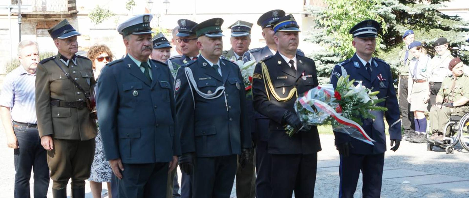 Grupa przedstawicieli służb mundurowych oczekuje na złożenie kwiatów przed pomnikiem żołnierzy 33 Pułku Piechoty