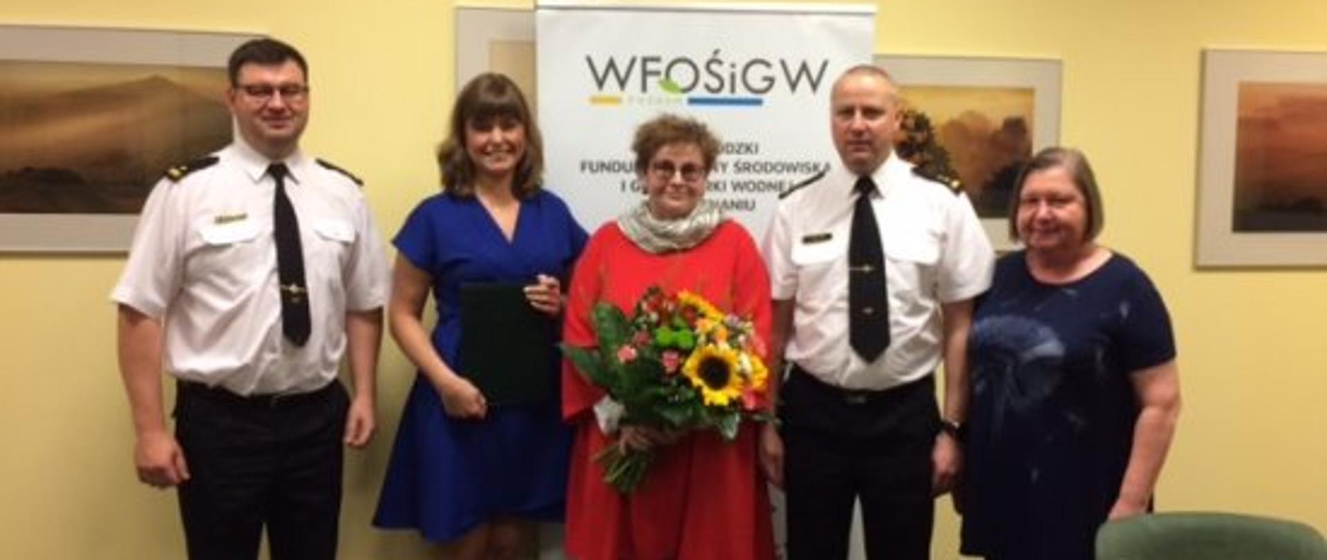 na tle żółtej ściany stoi grupa ludzi, z lewej strony stoi strażak ubrany w białą koszule z krawatem. Obok stoi kobieta w długich włosach w niebieskiej sukience, obok stoi kobieta w czerwonym swetrze z chustą na szyi i w okularach kobieta w środku trzyma bukiet kwiatów, obok sto strażak w białej koszuli z krawatem, obok stoi kobieta w niebieskiej koszuli