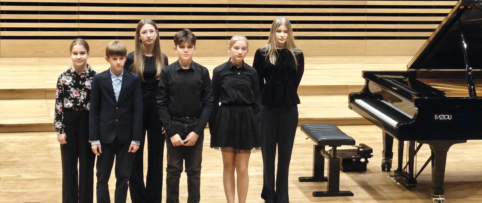 Wszyscy wykonawcy stoją na estradzie przy fortepianie.