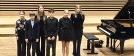 Wszyscy wykonawcy stoją na estradzie przy fortepianie.
