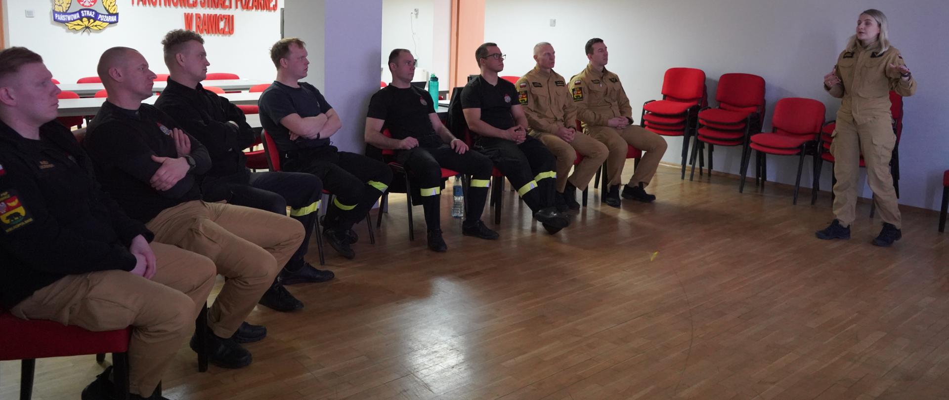 Świetlica komendy. Na krzesłach siedzą strażacy uczestniczący w zajęciach. Przed nimi stoi pani psycholog, która prowadzi psychoedukację. Wszyscy strażacy są umundurowani. W tle na ścianie widoczne jest logo PSP, nazwa komendy i drzwi ewakuacyjne. Z prawej strony, przy ścianie ustawione są czerwone krzesełka.