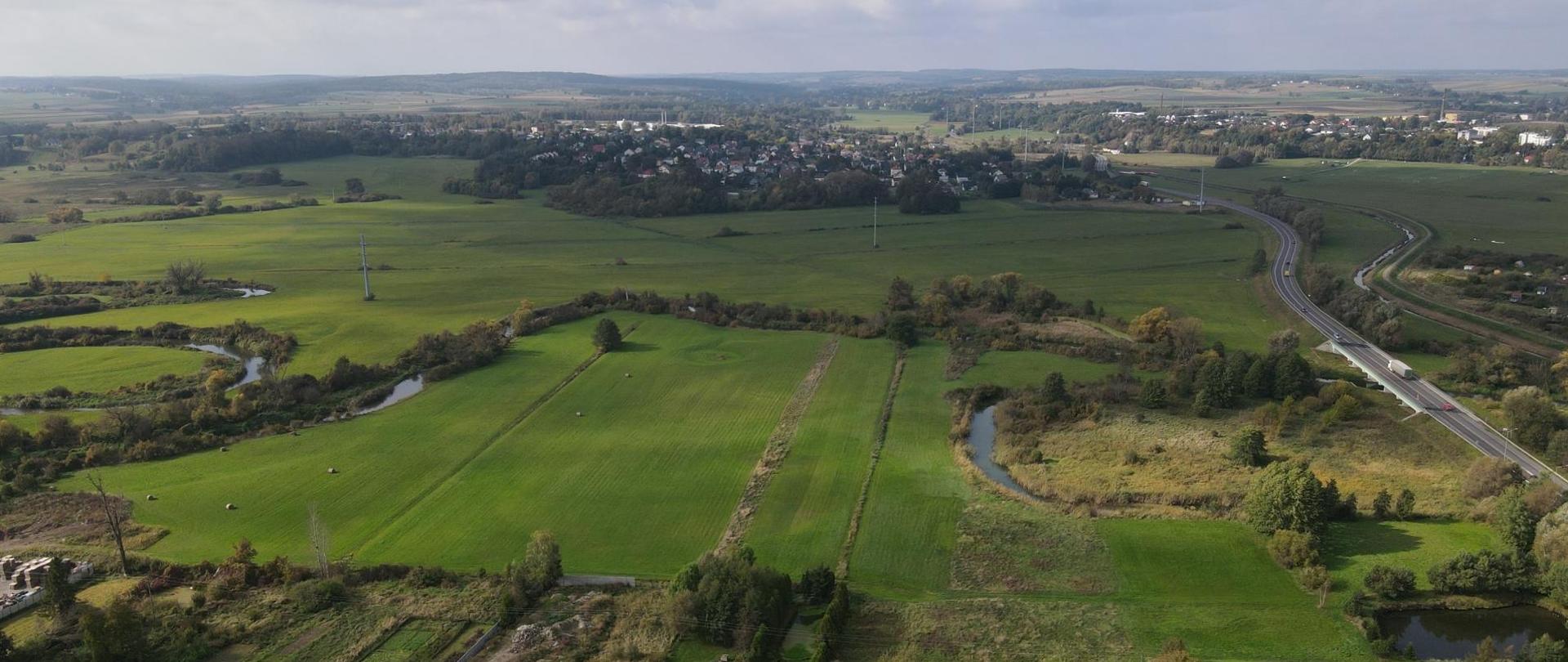 Na zdjęciu widoczny jest krajobraz wiejski z lotu ptaka. Widać zielone łąki. W okolicy znajdują się budynki mieszkalne o czerwonych i szarych dachach, otoczone drzewami i krzewami. Kręta droga przecina część terenu, a na niej widoczne są pojazdy. Dominuje czyste niebo z rozproszonymi chmurami, co wskazuje na dobre warunki pogodowe.