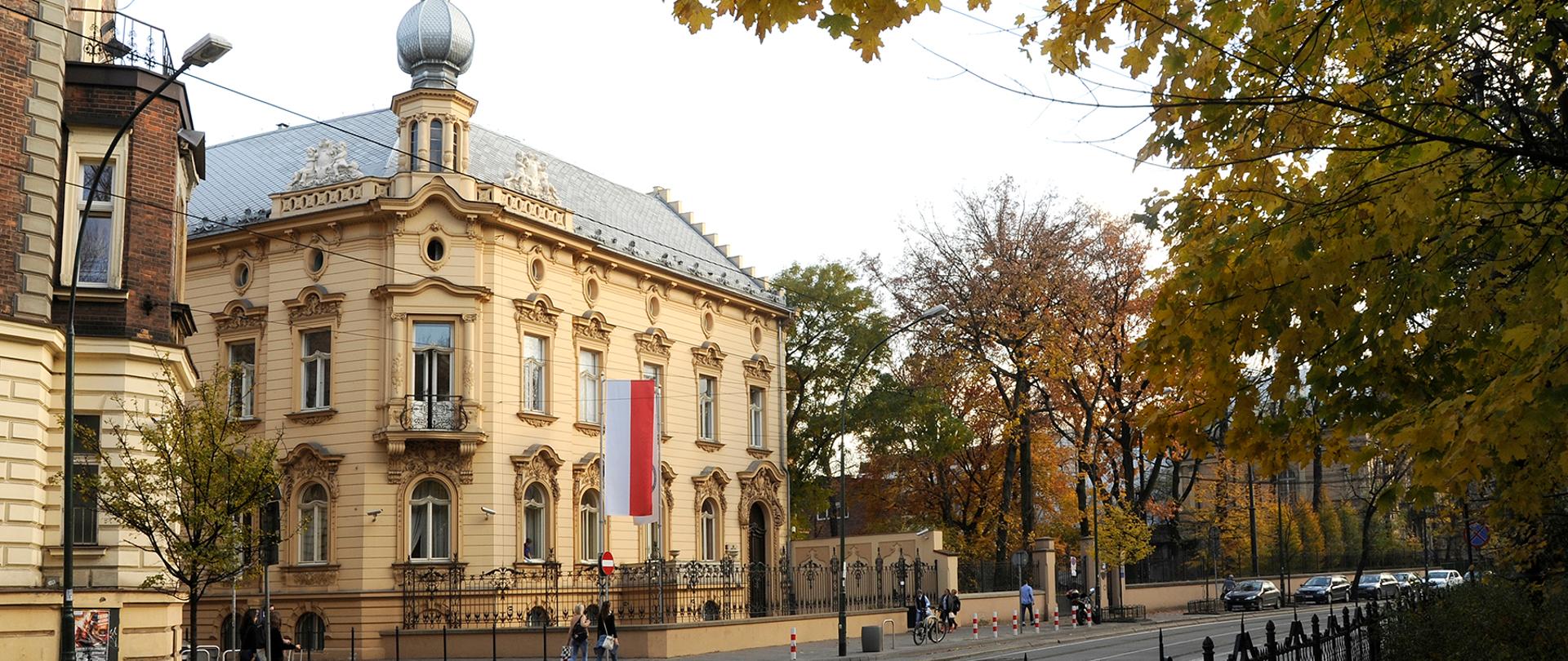 Oświetlona słońcem narożna fasada budynku instytutu Ekspertyz sądowych widziana z drugiej strony ulicy, na pierwszym planie drzewo z pożółkłymi jesiennymi liśćmi. 