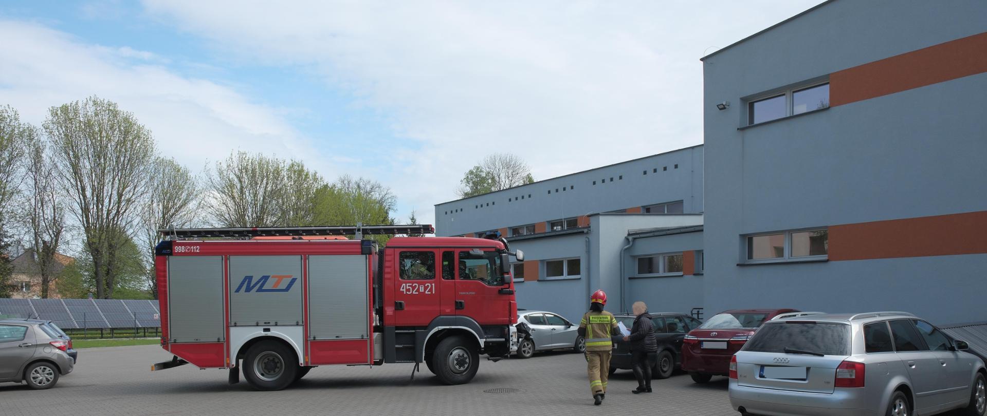 Na pierwszym planie wóz strażacki stojący bokiem pośród samochodów osobowych na parkingu za budynkiem szkoły.