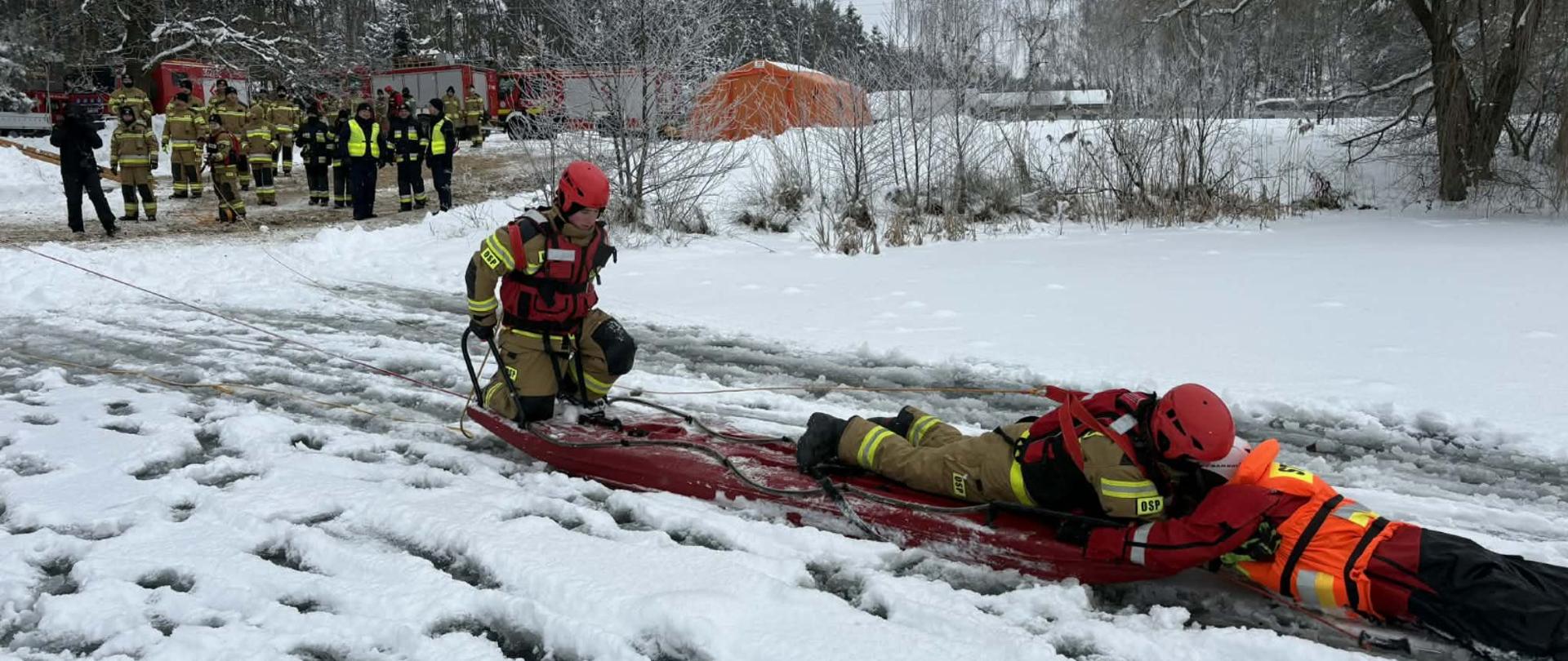 Strażacy ćwiczą na oblodzonym akwenie wodnym, widać strażaków na desce lodowej. W tle drzewa oraz zabudowania na brzegu stoją strażacy.