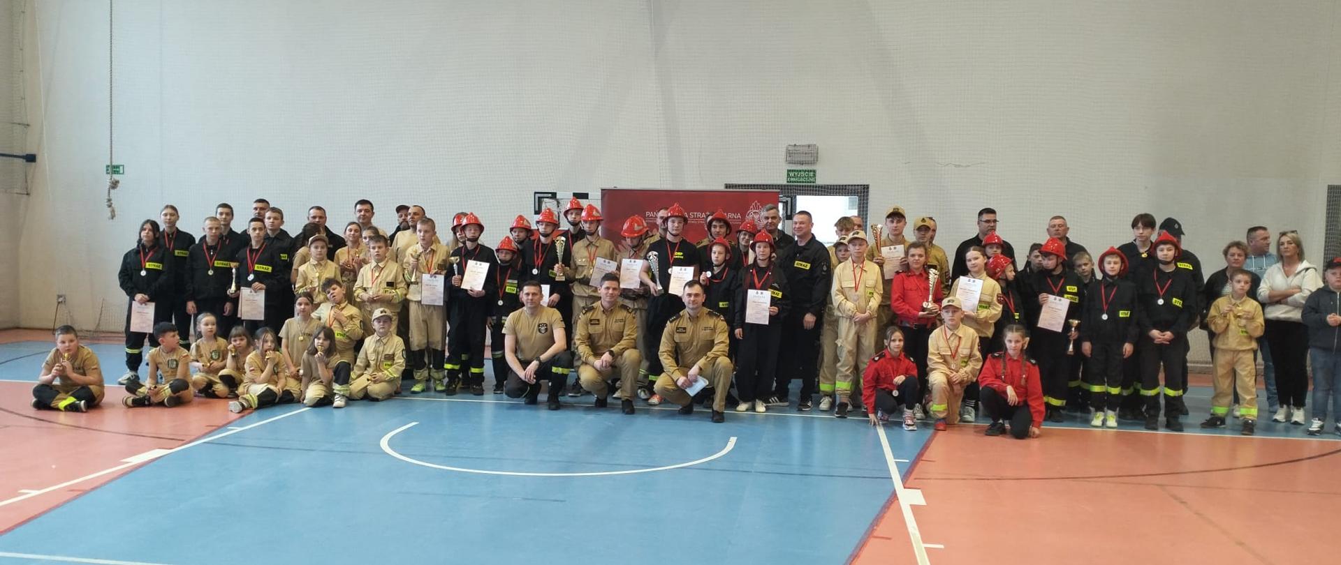 Szerokie ujęcie wykonane w hali sportowej, przedstawiające dużą grupę osób uczestniczących w wydarzeniu strażackim. Na środku, na tle jasnej ściany, stoi kilkudziesięciu uczestników w różnym wieku – od dzieci po dorosłych.
Większość osób ma na sobie mundury strażackie: Część grupy (głównie dorośli i młodzież) ubrana jest w ciemne, granatowe lub czarne mundury bojowe z odblaskowymi pasami. Dzieci i niektórzy opiekunowie noszą piaskowe (beżowe) mundury koszarowe. Wielu uczestników ma na głowach charakterystyczne czerwone hełmy strażackie.
Osoby w pierwszym rzędzie klęczą, pozostali stoją za nimi. W centralnej części widać osoby trzymające pamiątkowe dyplomy oraz puchary, co sugeruje zakończenie zawodów lub uroczystą zbiórkę. W tle, za uczestnikami, znajduje się czerwona tablica informacyjna lub ścianka reklamowa. Podłoga hali jest dwukolorowa: niebieskie pole do gry wyznaczone białymi liniami oraz pomarańczowa nawierzchnia wokół niego. Oświetlenie jest jasne i równomierne, typowe dla obiektów sportowych.