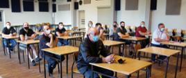 Widok z półprofilu. Na pierwszym planie za stołami na sali GOKiP siedzą uczestnicy spotkania – członkowie OSP - w maseczkach ochronnych. 