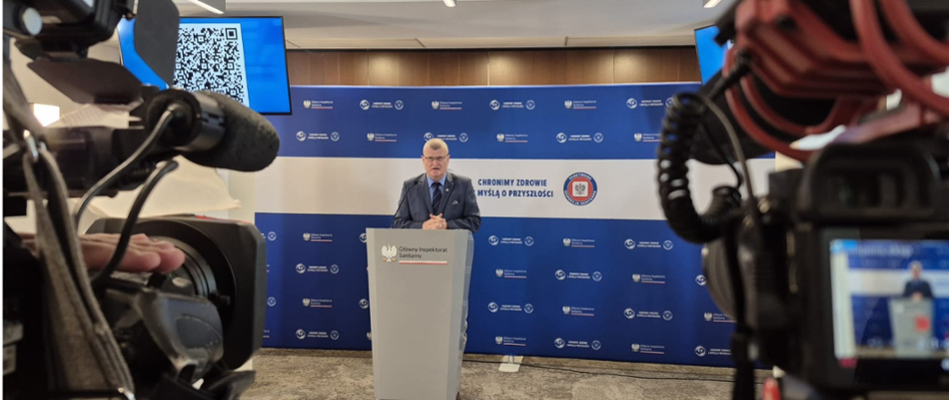 Paweł Grzesiowski przemawia przy mównicy Głównego Inspektoratu Sanitarnego podczas briefingu prasowego. W kadrze widoczne są kamery telewizyjne rejestrujące wydarzenie oraz tło z logo GIS i hasłem