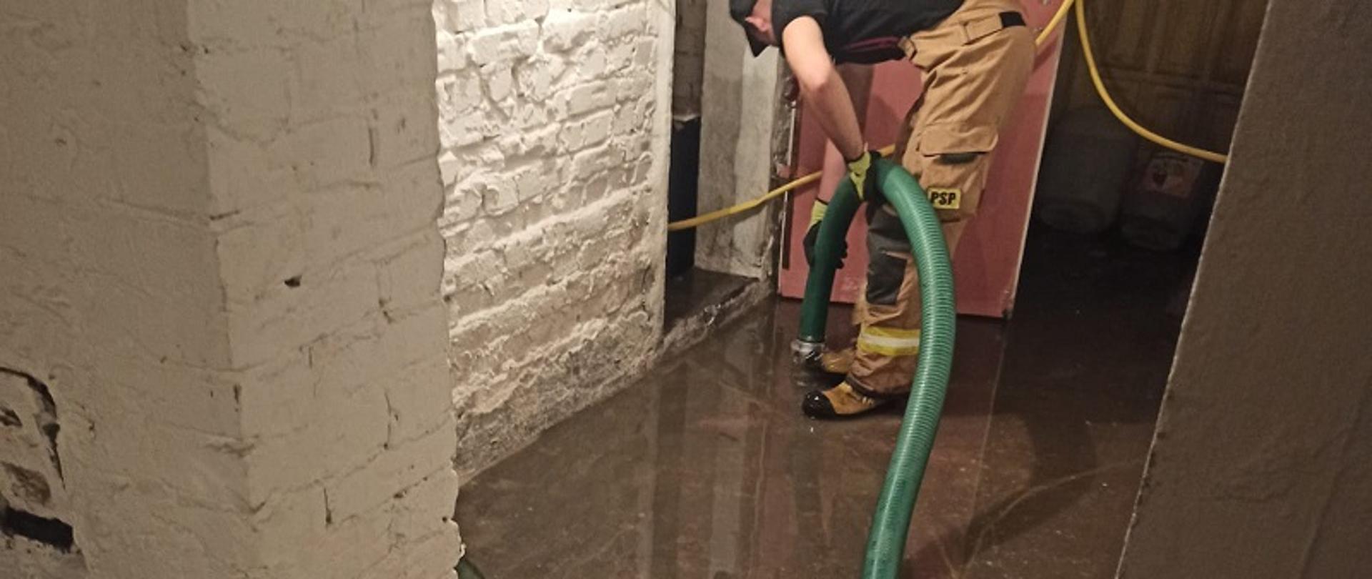 Zdjęcie przedstawia strażaka obsługującego pompę pływającą w zalanej piwnicy