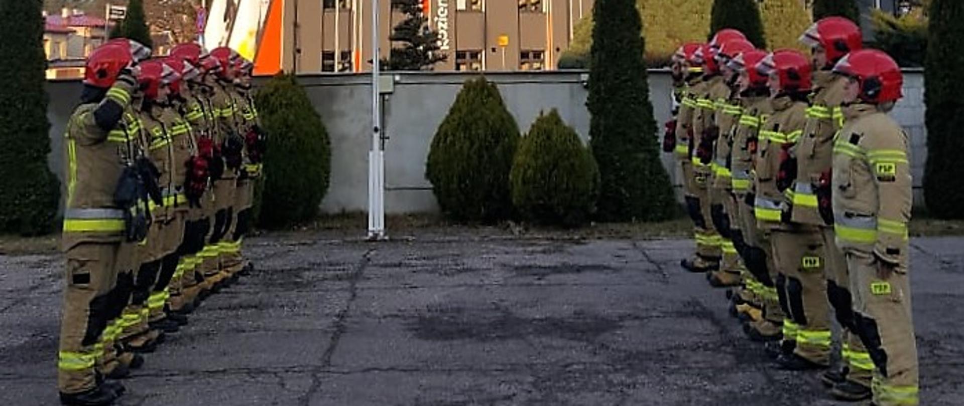 Strażacy w umundurowaniu specjalnym stoją w szeregach na przeciwko siebie dokonując uroczystej zmiany służby, za nimi widać masz z podniesioną flagą Polski.