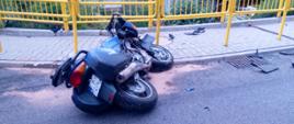 Zdjęcie przedstawia leżący na boku motocykl. W tle widać żółte barierki znajdujące się na chodniku.