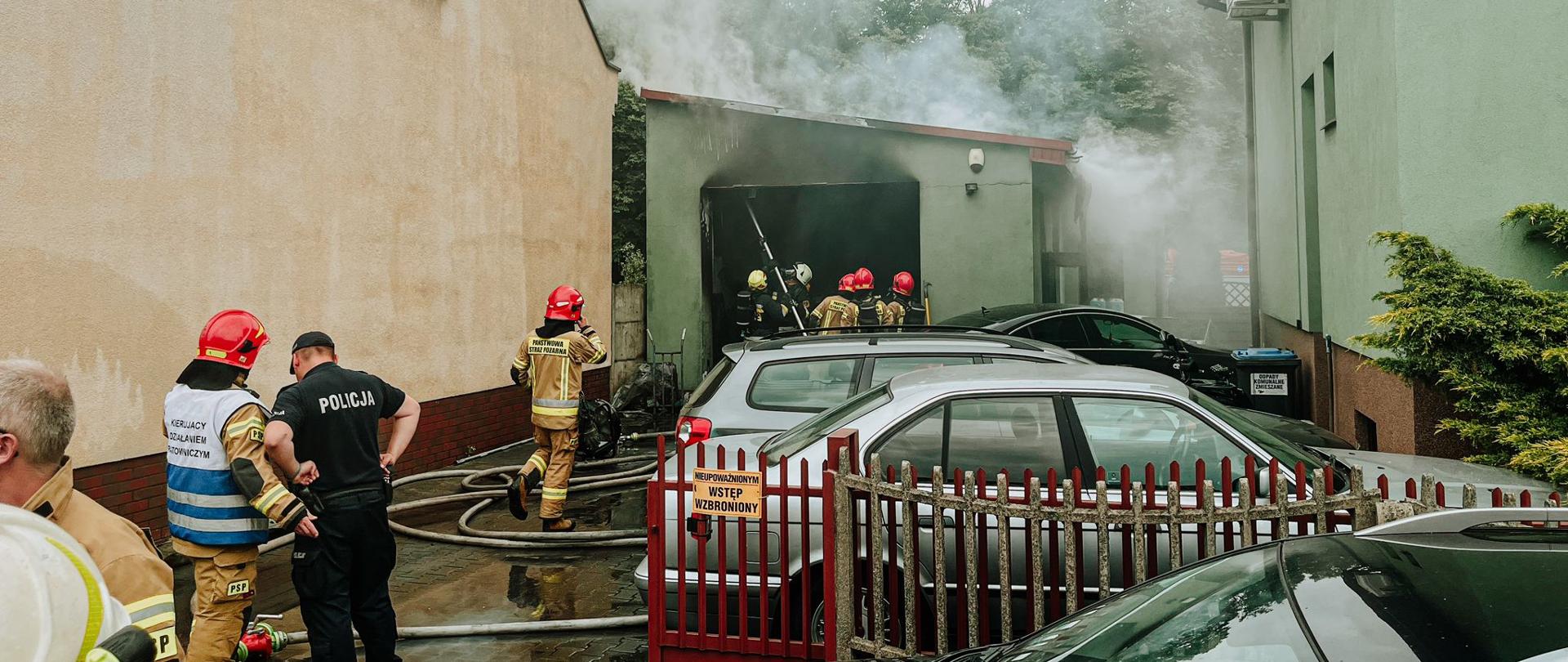 Na dalszym planie strażacy w trakcie działań gaśniczych w warsztacie. Z budynku wydobywa się dym. Bliżej stoją samochody zaparkowane bokiem na posesji, po lewej stronie budynek mieszkalny. Przy budynku stoją strażacy i policjant.