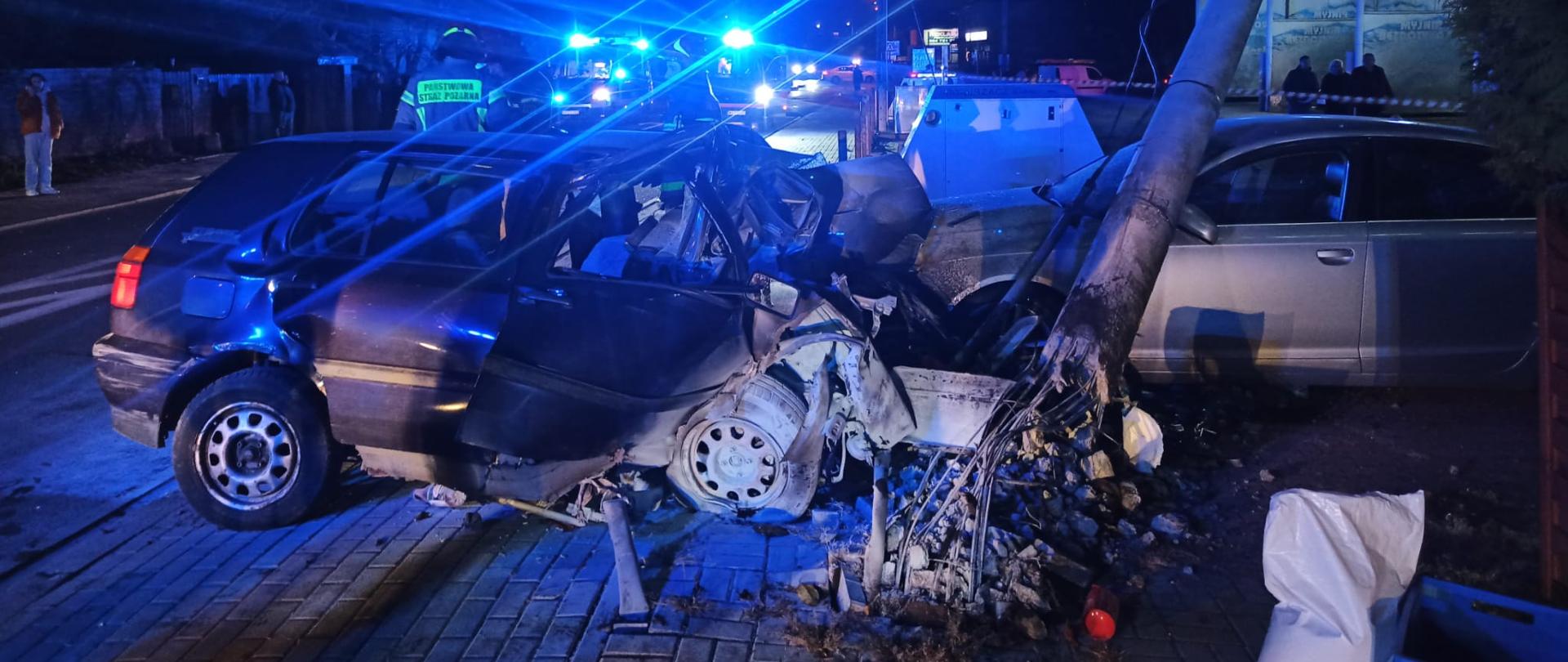 Fotografia w centralnym punkcie przedstawia pojazd, który bezpośrednio uczestniczył w zdarzeniu i doprowadził do uszkodzenia słupa. Na pierwszym planie można zauważyć słup, który w skutek zdarzenia został przewrócony na prawidłowo zaparkowany srebrny pojazd. W dalszej części zdjęcia widzimy akcję ratowniczą prowadzona przez strażaków oraz Zespół Ratownictwa Medycznego. 