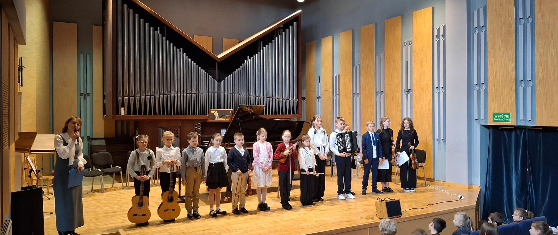 Na zdjęciu widać grupę dzieci i młodzieży stojącą na scenie w sali koncertowej, prawdopodobnie po występie muzycznym. Oto główne elementy fotografii: Wykonawcy: Na scenie stoi kilkanaścioro dzieci w strojach galowych. Niektóre z nich trzymają instrumenty, m.in. gitary klasyczne, skrzypce oraz akordeon. Po lewej stronie stoi dorosła kobieta z mikrofonem, która prawdopodobnie prowadzi wydarzenie. Instrumenty w tle: Centralnym punktem tła są imponujące, nowoczesne organy piszczałkowe o charakterystycznym kształcie litery „V”. Przed nimi znajduje się czarny fortepian skrzydłowy. Wnętrze: Sala ma nowoczesny wystrój z dużą ilością jasnego drewna na ścianach i podłodze. Po prawej stronie widoczne jest zielone oznakowanie wyjścia ewakuacyjnego w języku polskim („Wyjście ewakuacyjne”). Publiczność: W dolnej części zdjęcia widać tyły głów widzów siedzących w niebieskich fotelach, co sugeruje, że jest to szkolny popis lub oficjalny koncert.