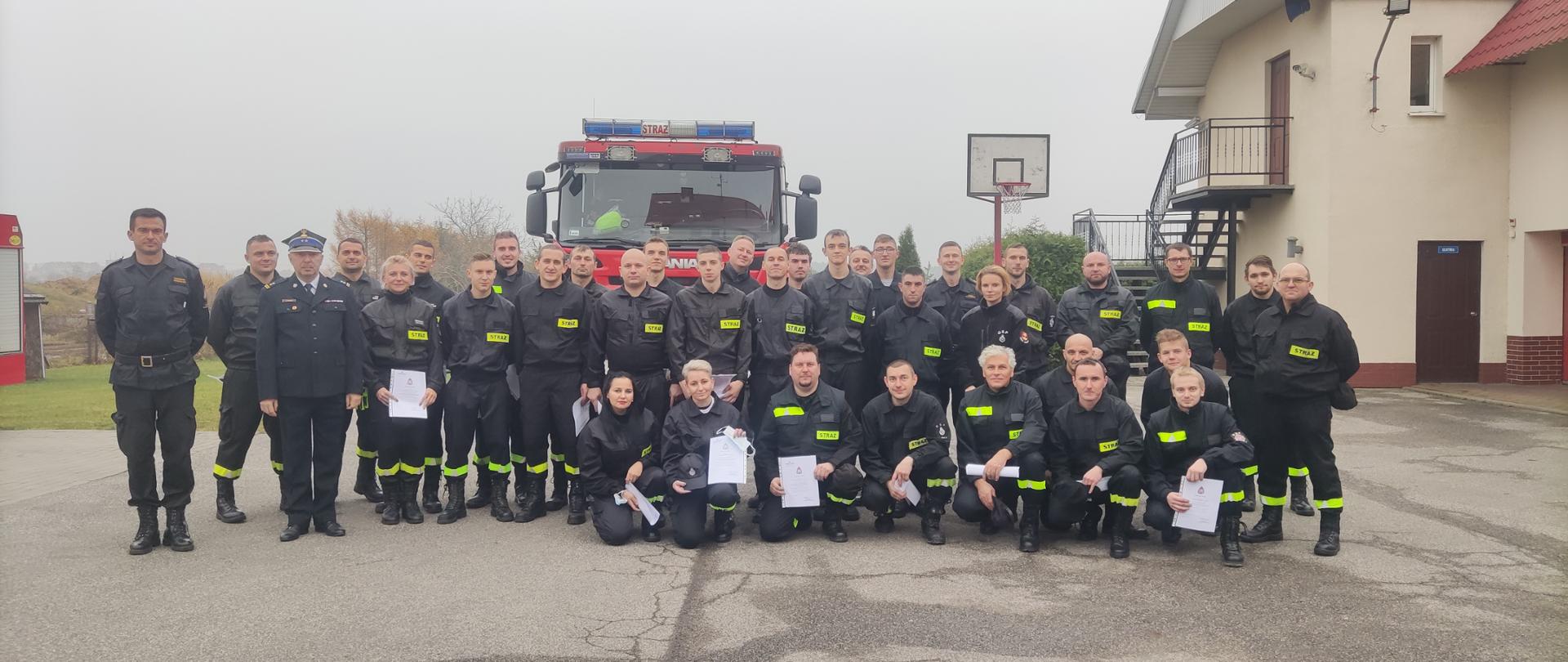 Zdjęcie przedstawia teren Komendy Powiatowej Państwowej Straży Pożarnej w Radziejowie, strażaków PSP oraz OSP na tle pojazdu pożarniczego podczas zakończenia szkolenia podstawowego strażaków ratowników Ochotniczych Straży Pożarnych.