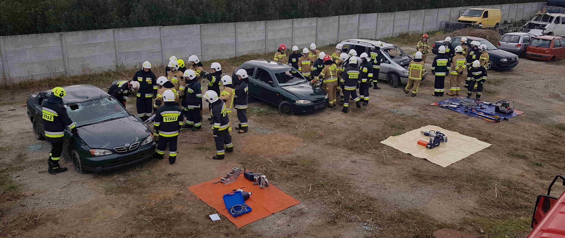Zdjęcie zrobione w dzień na auto złomie. Na zdjęciu widać cztery grupy strażaków podczas pracy narzędziami hydraulicznymi. Strażacy tną samochody osobowe. 