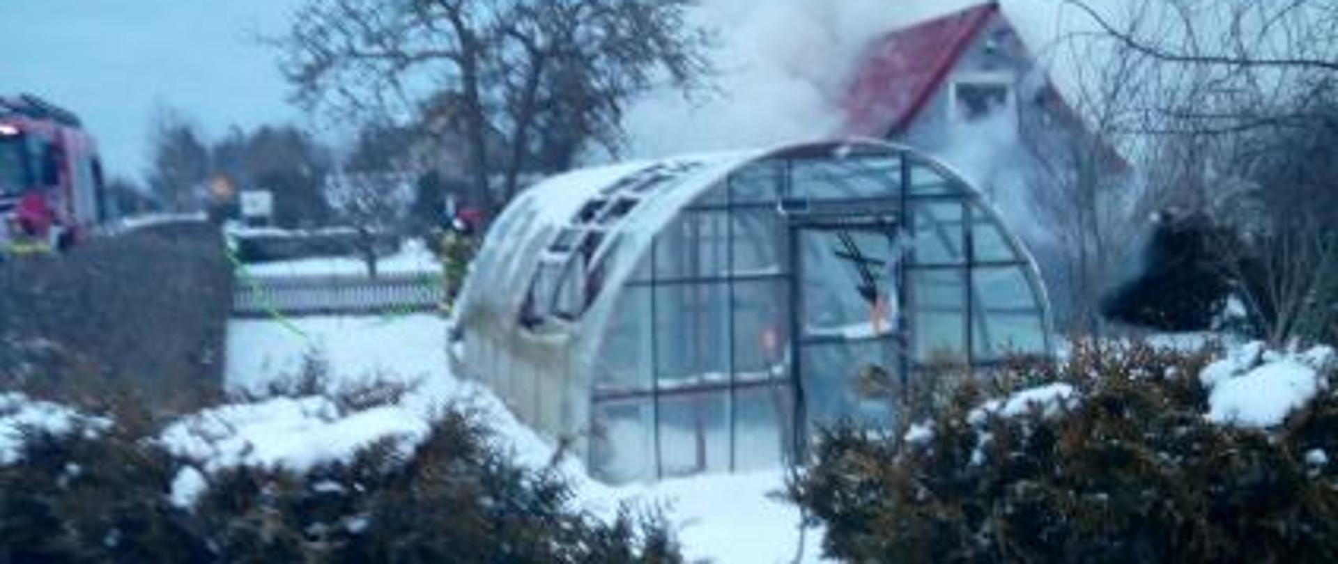 Altana ogrodowa objęta pożarem – widoczny dym unoszący się nad konstrukcją w zimowej scenerii.