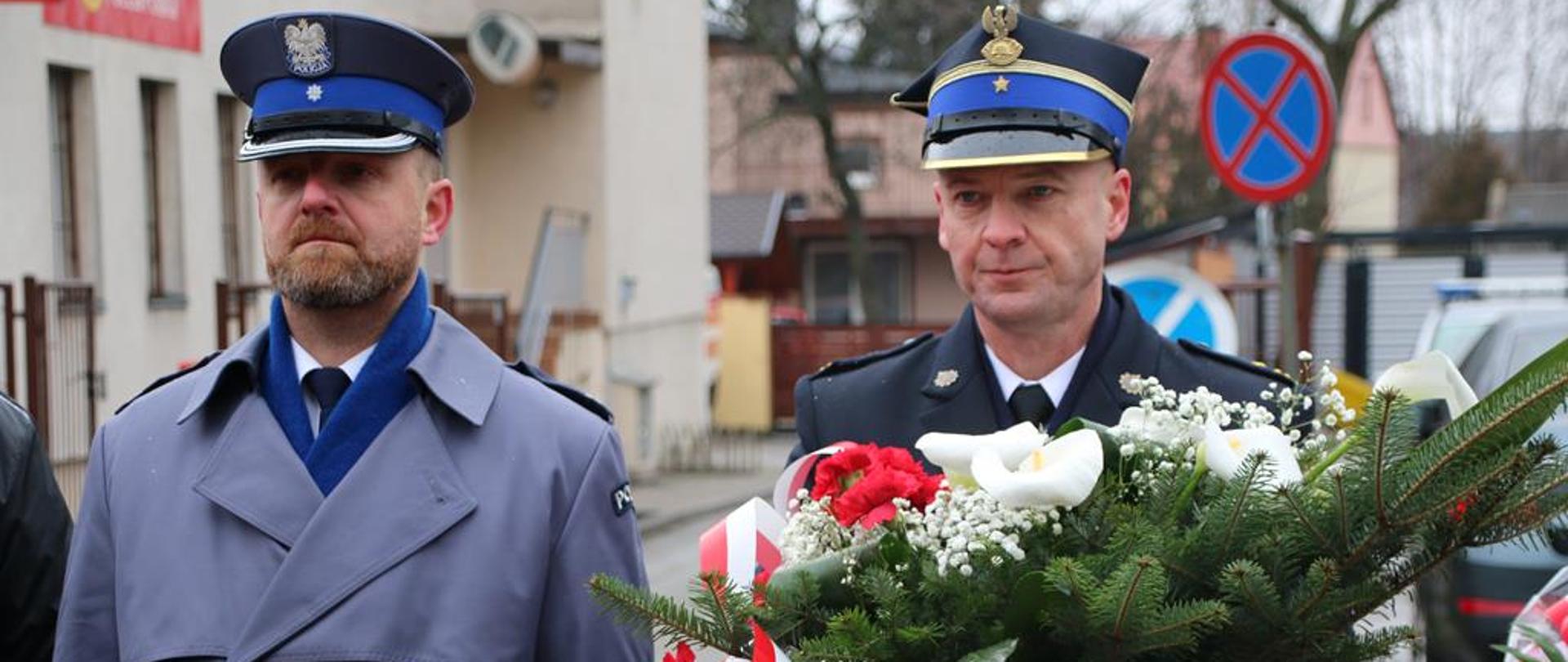 Na pierwszym palnie widnieje postać Komendanta Powiatowego Państwowej Straży Pożarnej w Jędrzejowie w umundurowaniu galowym oraz płaszczu. Na głowie ma założoną czapkę rogatywkę. W rękach trzyma wiązankę biało- czerwonych kwiatów. Po jego lewej stronie znajduje się zastępca komendanta policji także w umundurowaniu galowym oraz płaszczu.
