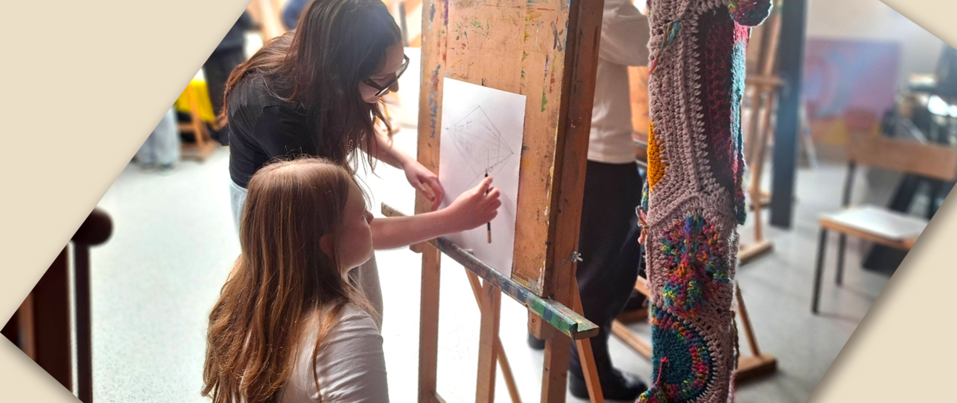 Przy szta Lutze siedzi uczennica szkoły podstawowej, starsza koleżanka z liceum prezentuje jej jak rysować perspektywę; po prawej stronie Słup drewniany owinięty kolorową robót K na szydełku, w tle pracownia malarstwa