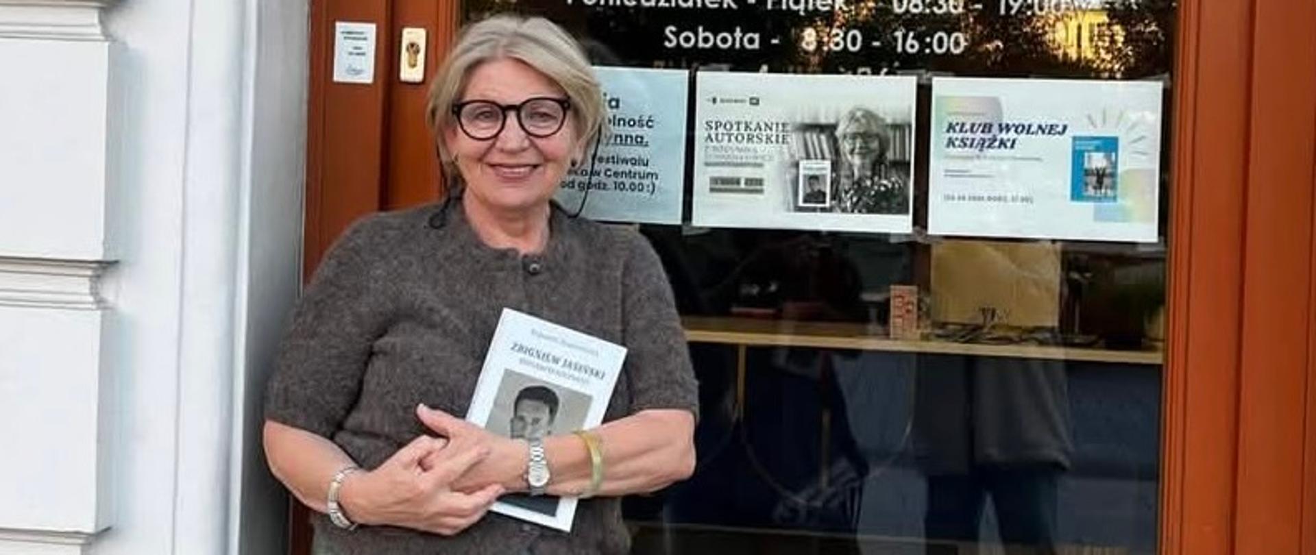 The writer is standing in front of a bookstore door, holding a book in her hand.