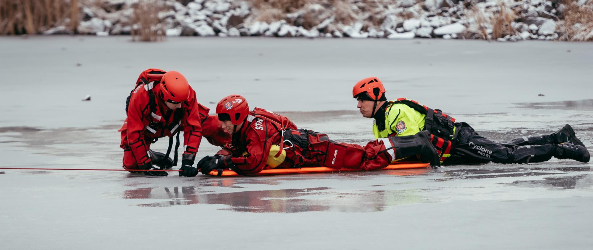 Strażacy w kombinezonach wodnych podejmują poszkodowanego z przerębla na deskę ortopedyczną którą po tafli lodu ciągną liną aby dostać się na brzeg