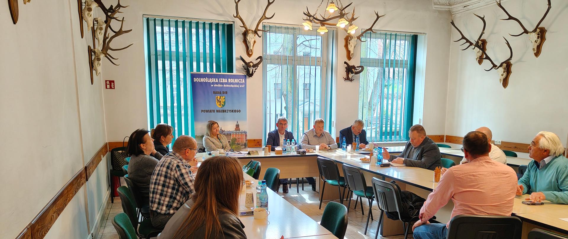 Spotkanie kilkunastu osób w sali konferencyjnej – uczestnicy siedzą przy stołach w układzie podkowy, słuchając osób prowadzących przy stole głównym. Na ścianach widoczne są poroża zwierząt, a w tle stoi baner organizacji.