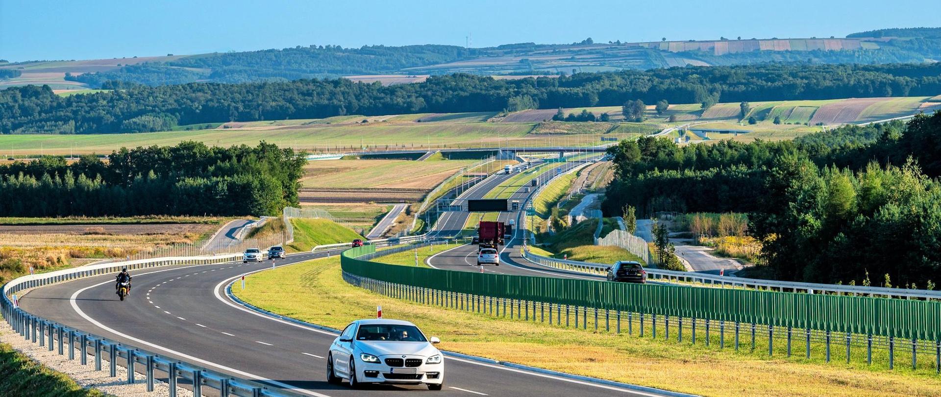 Droga ekspresowa S7 na odcinku Moczydło - Miechów. Dwujezdniowa droga ekspresowa wijąca się między polami i lasami po niewielkich pagórkach. Widoczne pojazdy poruszające się w obu kierunkach.