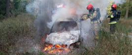 Zdjęcie przedstawia palący się samochodu osobowego typu kombi. Palące się auto znajduję się na poboczu drogi leśnej. Auto pokrywane jest pianą gaśniczą przez dwóch strażaków ubranych w ubrania specjalne w sprzęcie ochrony układu oddechowego. Za palącym autem znajduje się wóz strażacki.