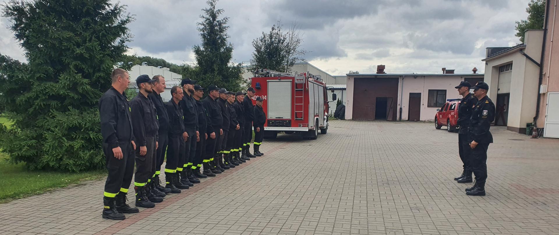 Zdjęcie przedstawia zbiórkę strażaków i druhów, wykonane w dniu egzaminu kończącego szkolenie kierowców-konserwatorów Ochotniczej Straży Pożarnej. Na zdjęciu widać kilkunastu druhów ustawionych w szeregu. Około czterech metrów na przeciwko stoją dwaj funkcjonariusze Państwowej Straży Pożarnej, Komendant Powiatowy oraz dowódca Jednostki Ratowniczo-Gaśniczej nr 1 w Aleksandrowie Kujawskim. Druhowie są ubrani w ubranie koszarowe, a strażacy w ubranie dowódczo-sztabowe. Zbiórka odbywa się obok jednostki, na placu pokrytym kostką brukową. W tle widać samochód ratowniczo-gaśniczy, samochód operacyjny, garaż oraz drzewa.