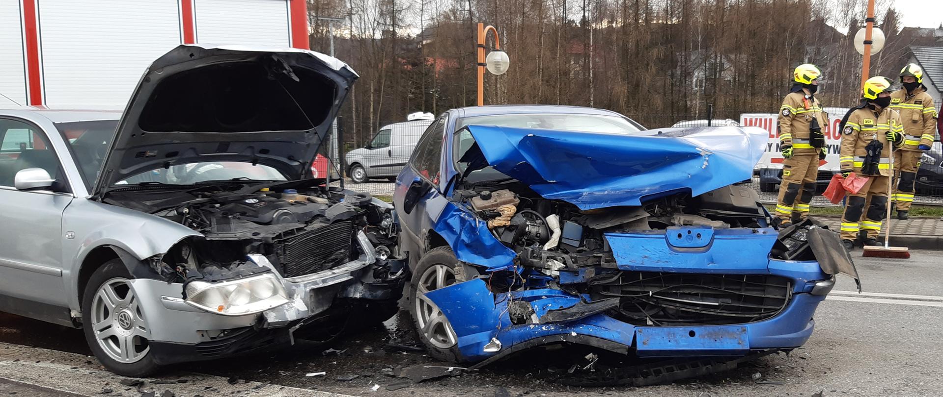 Na zdjęciu dwa samochody osobowe po zderzeniu czołowym z widocznymi zniszczeniami. Na drugim planie trzech strażaków oraz samochód ratowniczo gaśniczy