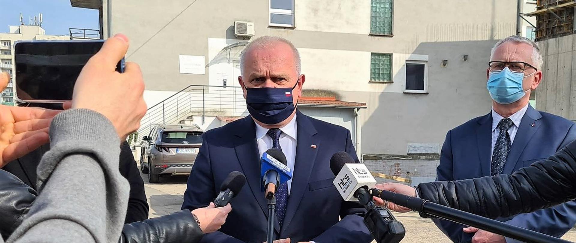 Wojewoda z Prezesem zarządu szpitala w czasie konferencji prasowej. W tle budowa. Na pierwszym Planie dziennikarze z mikrofonami i telefonami. 