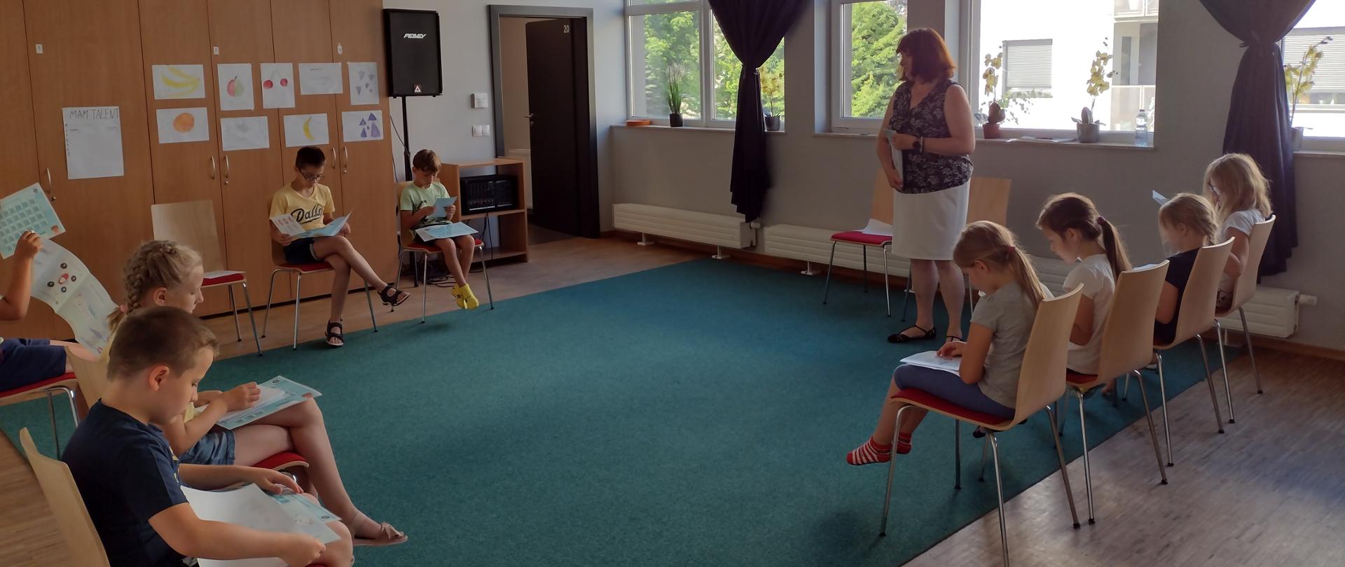 Na dużej sali dzieci siedzą w kręgu i ogladają rozdane przez prelegentkę ulotki.