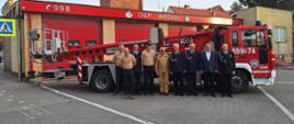 Plac przed remizą OSP Wronki. Na tle budynku oraz samochodu specjalnego z podnośnikiem hydraulicznym stoją uczestnicy uroczystości. Są wśród nich zarówno strażacy w ubraniach wyjściowych, koszarowych oraz służbowych w kolorze piaskowym, jak również osoba cywilna. 